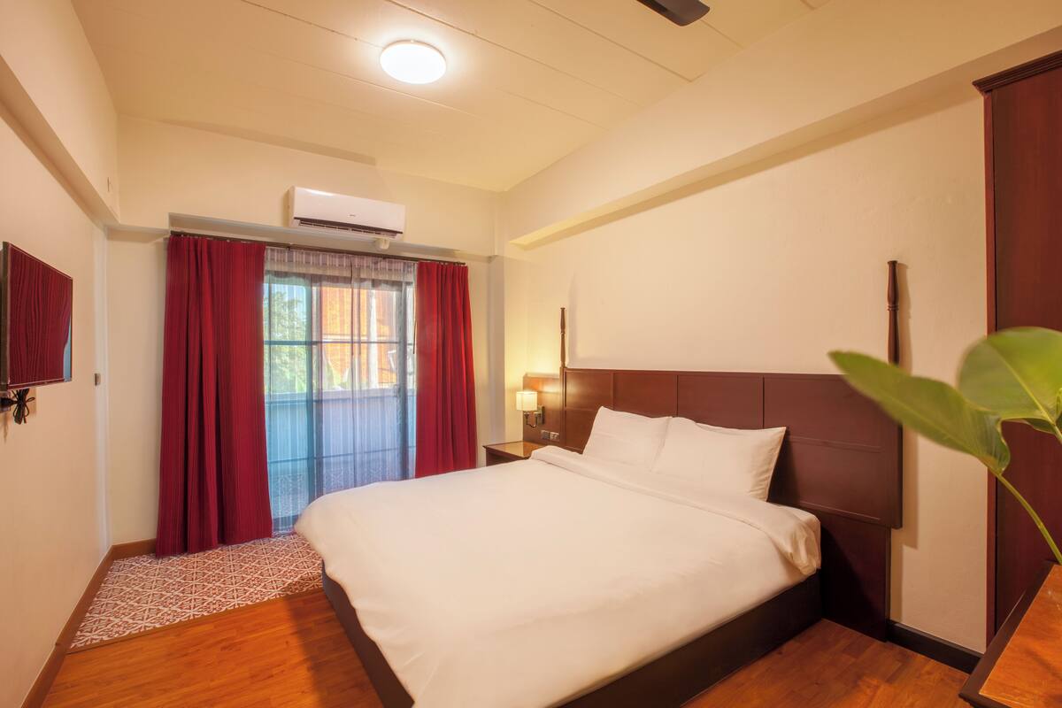 A hotel room with a large bed, a wooden floor, a window with red curtains, and an air conditioning unit on the wall.
