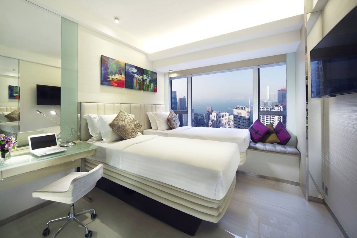 Habitación de hotel moderna con vistas a la ciudad, un escritorio con una computadora portátil y una cama grande con muchos almohadones.