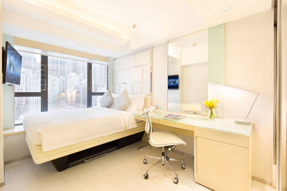 Habitación de hotel moderna con cama, escritorio, silla y ventanas amplias con vista a la ciudad.