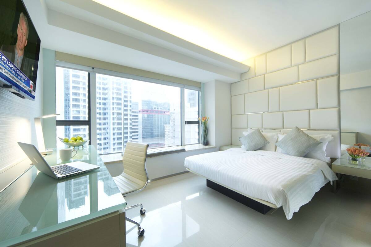Habitación de hotel moderna con una cama grande, un escritorio con una computadora portátil, una silla y vistas de la ciudad a través de ventanas amplias.