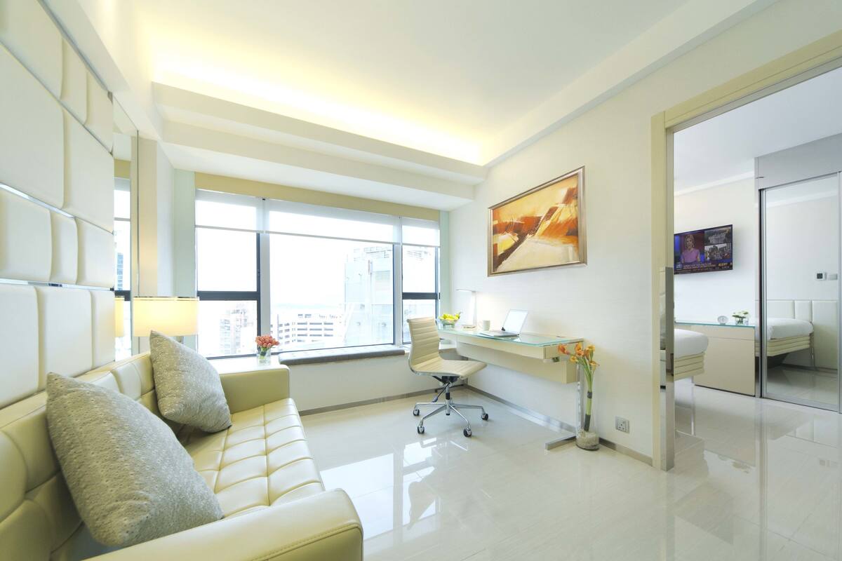 Una habitación de hotel moderna con un sofá de cuero blanco, un escritorio de vidrio y un ventanal con vistas a la ciudad.