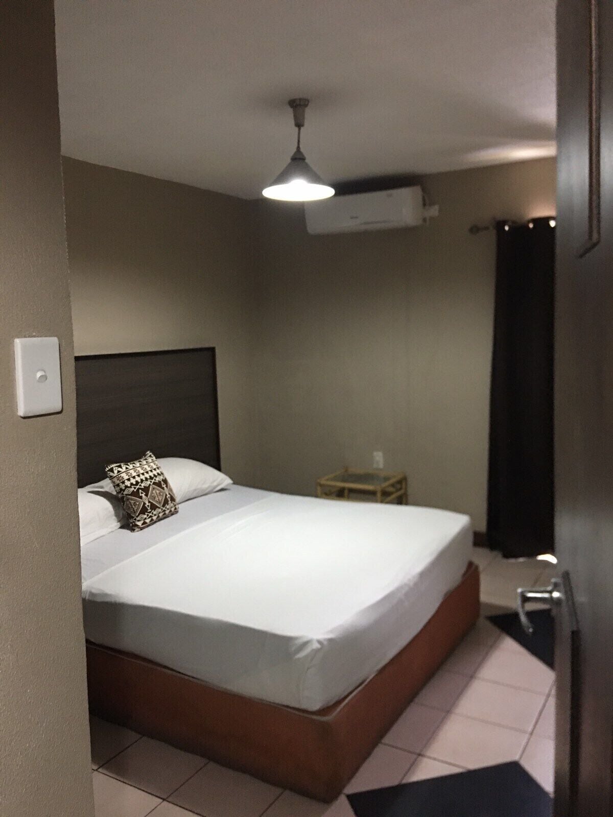 A bedroom with a bed, a bedside table, a lamp, and an air conditioning unit.