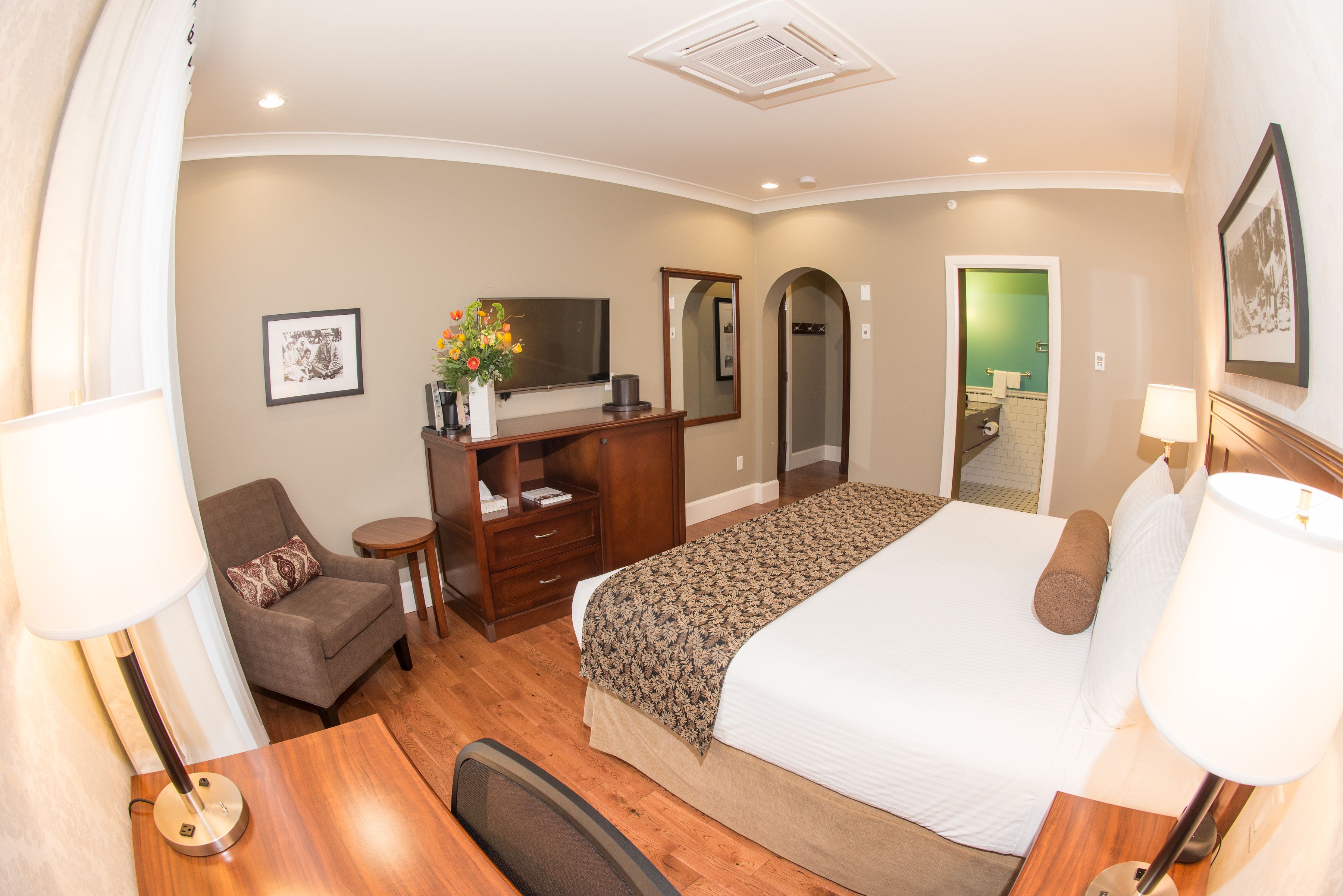 A hotel room with a large bed, a desk, a chair, a TV, a mirror, and a bathroom visible through an arched doorway.