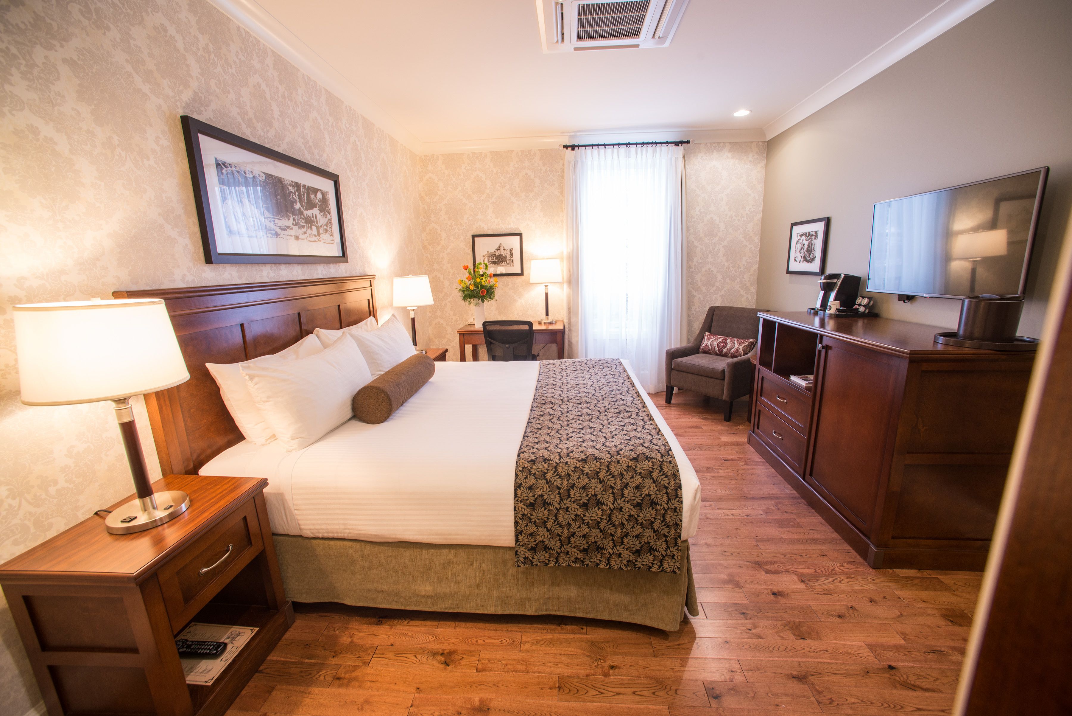 A hotel room with a large bed, bedside lamps, a desk with a chair, a television, and a framed picture on the wall.