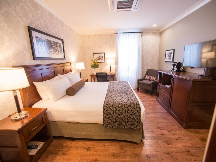 A hotel room with a large bed, bedside lamps, a desk with a chair, a television, and a framed picture on the wall.