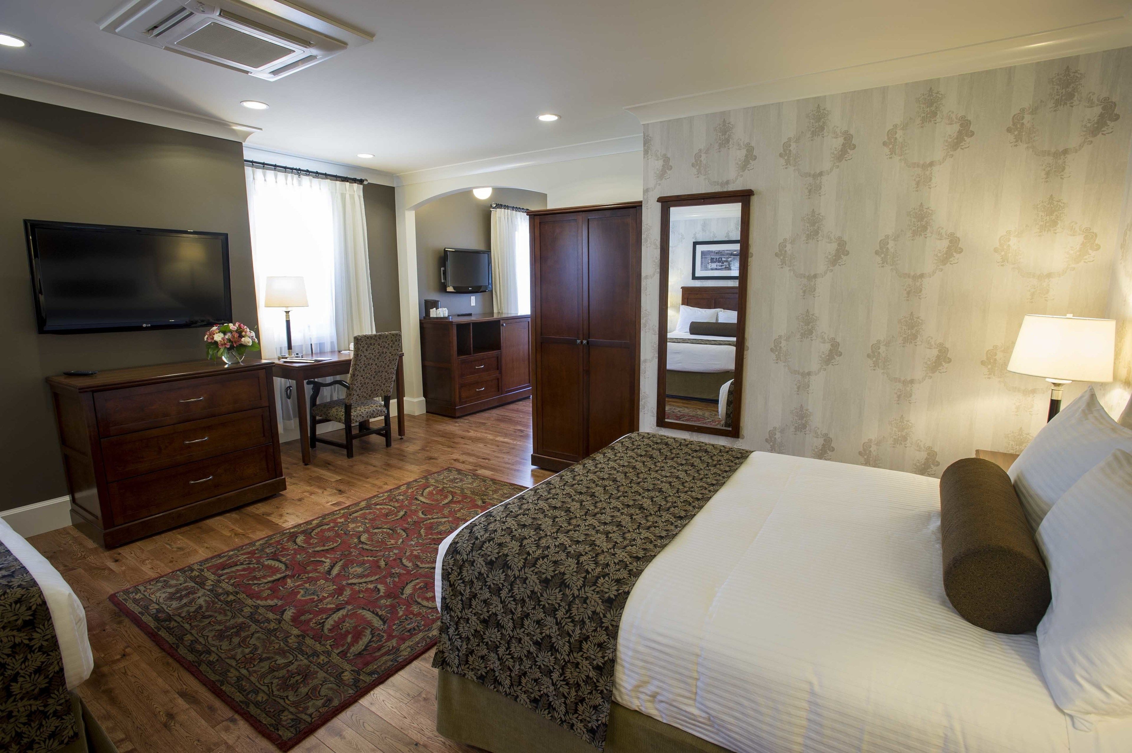 A hotel room with a bed, a desk with a chair, a television, a wardrobe, and a mirror.