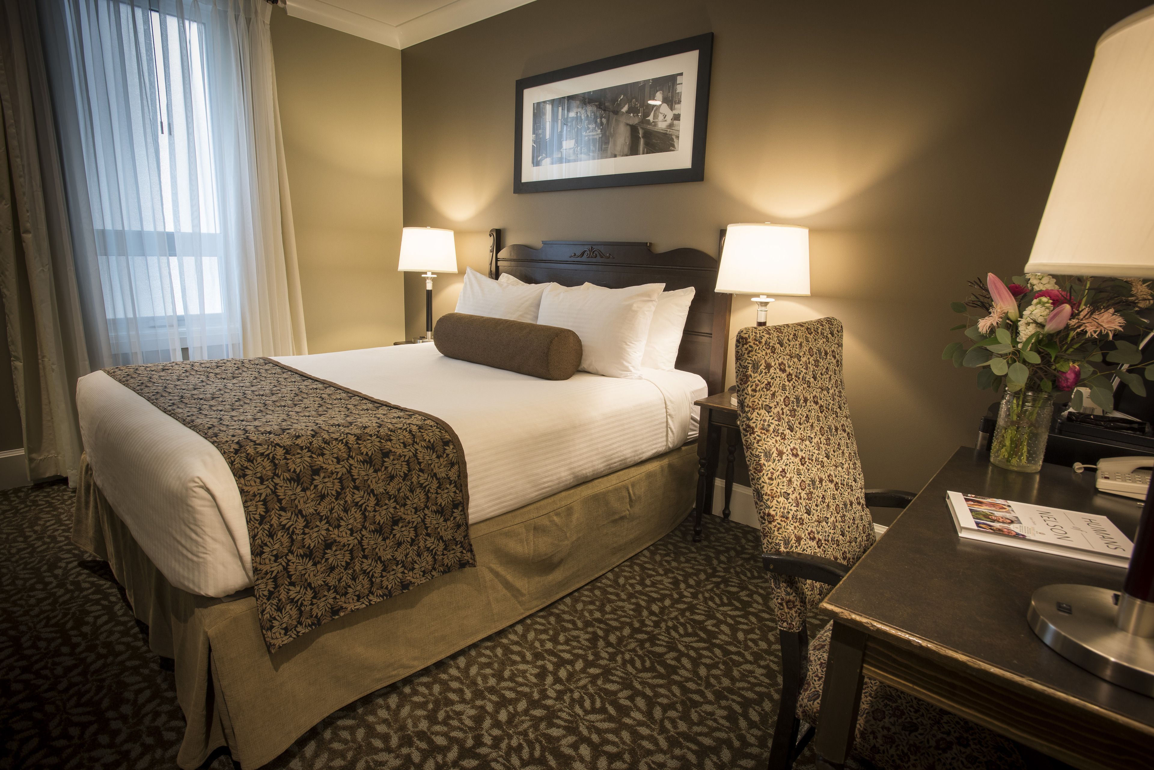A hotel room with a large bed, two bedside lamps, a chair, a desk with a lamp, and a framed picture on the wall.