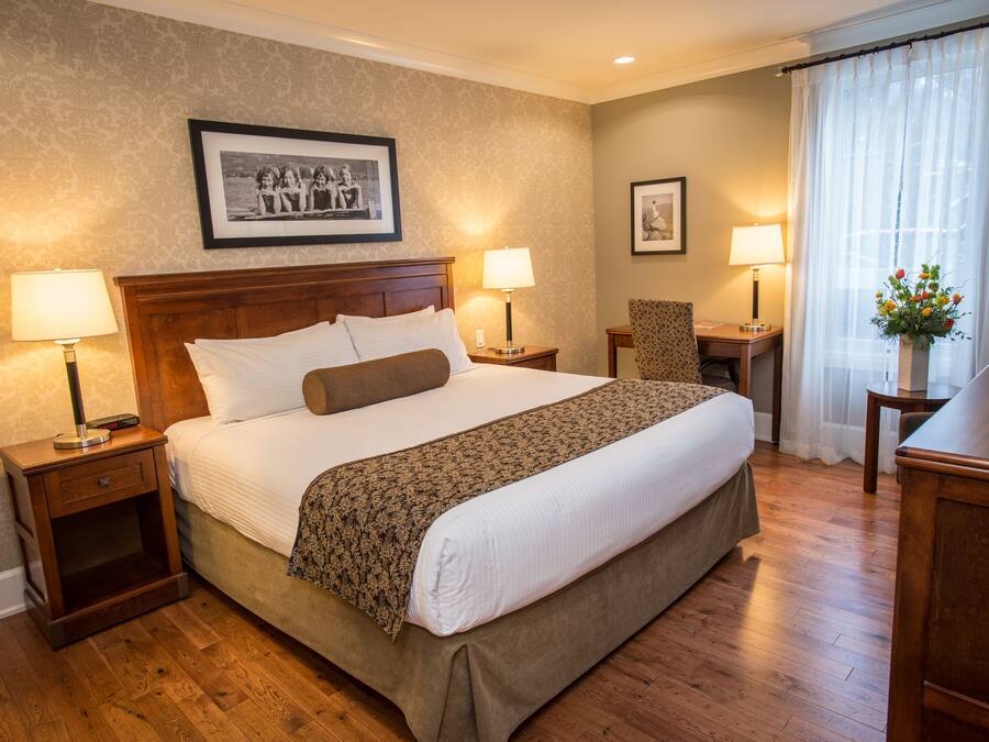 Une chambre d’hôtel avec un grand lit, deux tables de chevet avec des lampes, une chaise, un bureau et une fenêtre avec des rideaux.