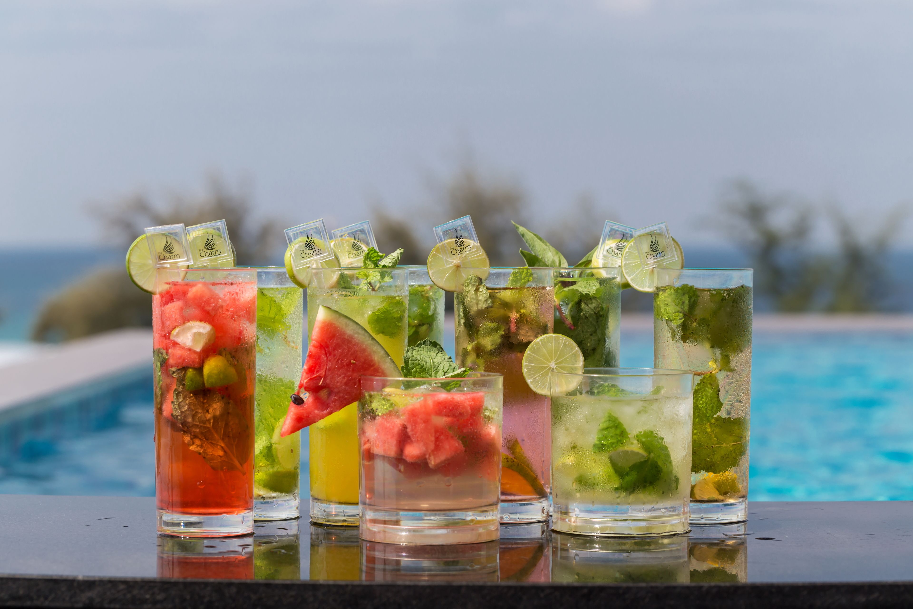 Une rangée de six grands verres remplis de diverses boissons, décorés de quartiers de citron vert et de feuilles de menthe, est placée sur une surface réfléchissante au bord d’une piscine, avec un arrière-plan naturel flou.