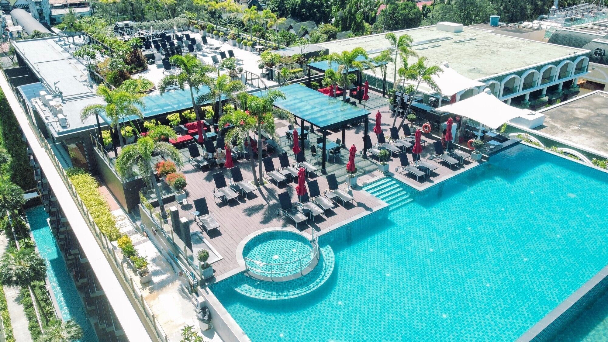 A rooftop pool area with lounge chairs and umbrellas, surrounded by greenery and a large building in the background.