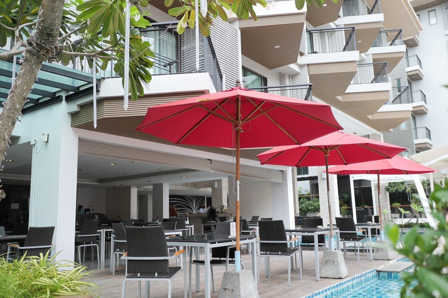 An outdoor seating area with red umbrellas, a pool, and a modern building with balconies.