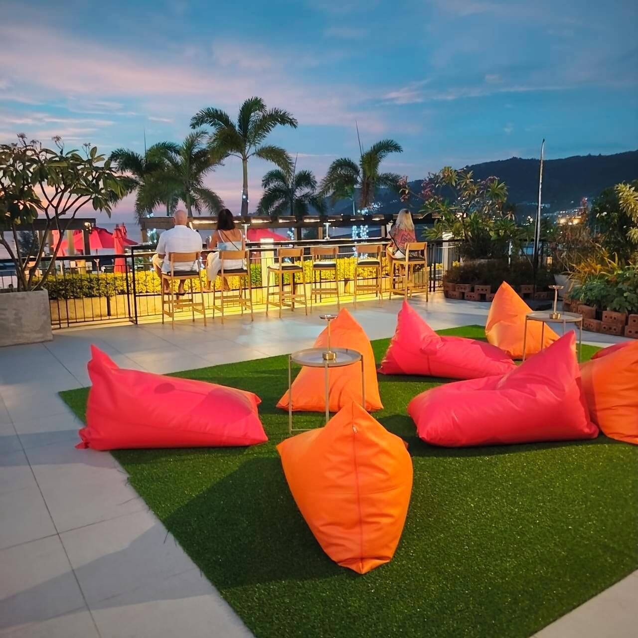 An outdoor seating area with orange bean bags and a small table, surrounded by palm trees and chairs, with a view of the mountains in the background.