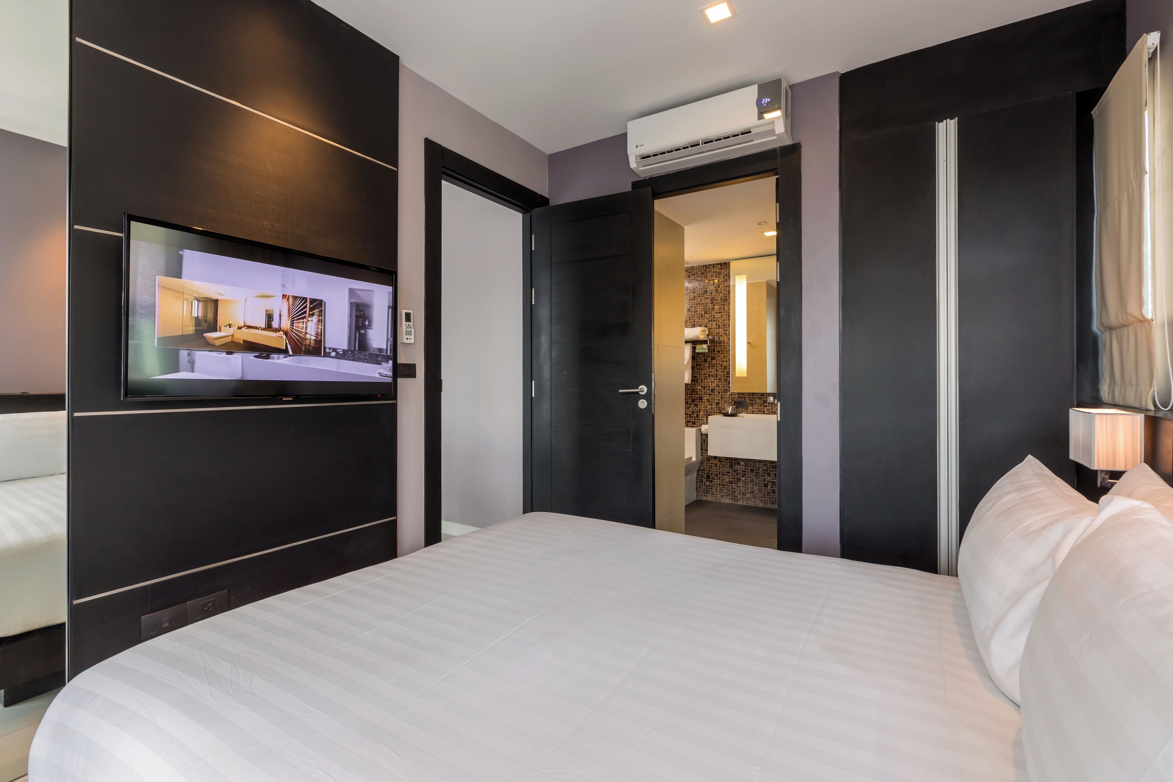 A modern hotel room with a large bed, a flat-screen TV mounted on the wall, a bathroom with a tiled shower, and an air conditioning unit.