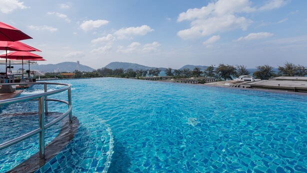 Una piscina azul cristalina con barandilla metálica, sombrillas rojas y vistas a montañas al fondo.