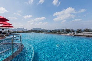 A clear blue pool with a metal railing, red umbrellas, and a view of mountains in the background. - The Charm Resort Phuket (Patong)