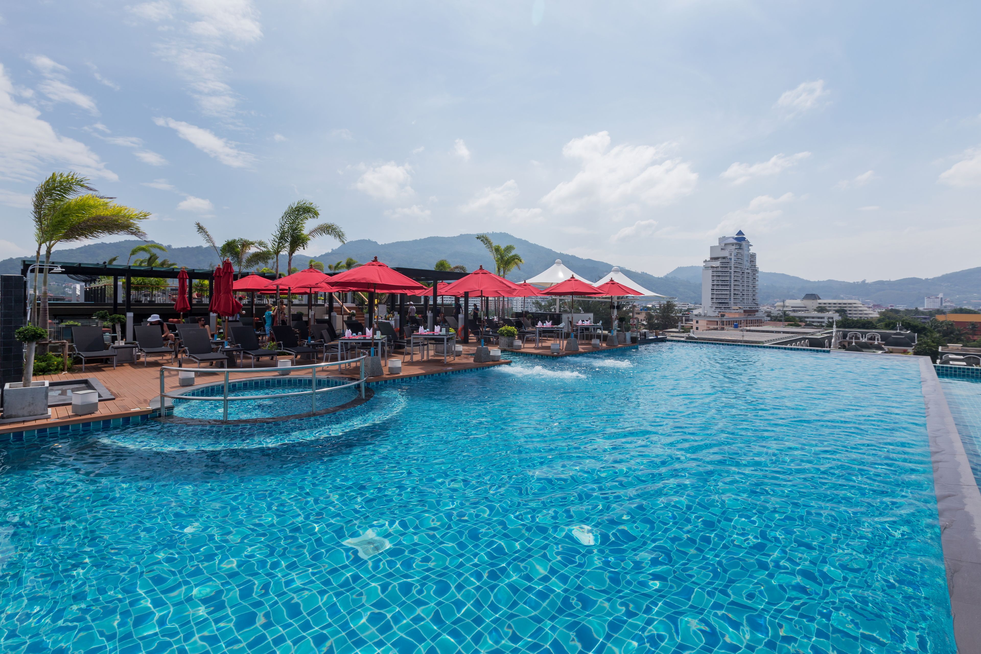 A swimming pool with red umbrellas and lounge chairs, surrounded by buildings and mountains.