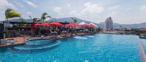 A swimming pool with red umbrellas and lounge chairs, surrounded by buildings and mountains.