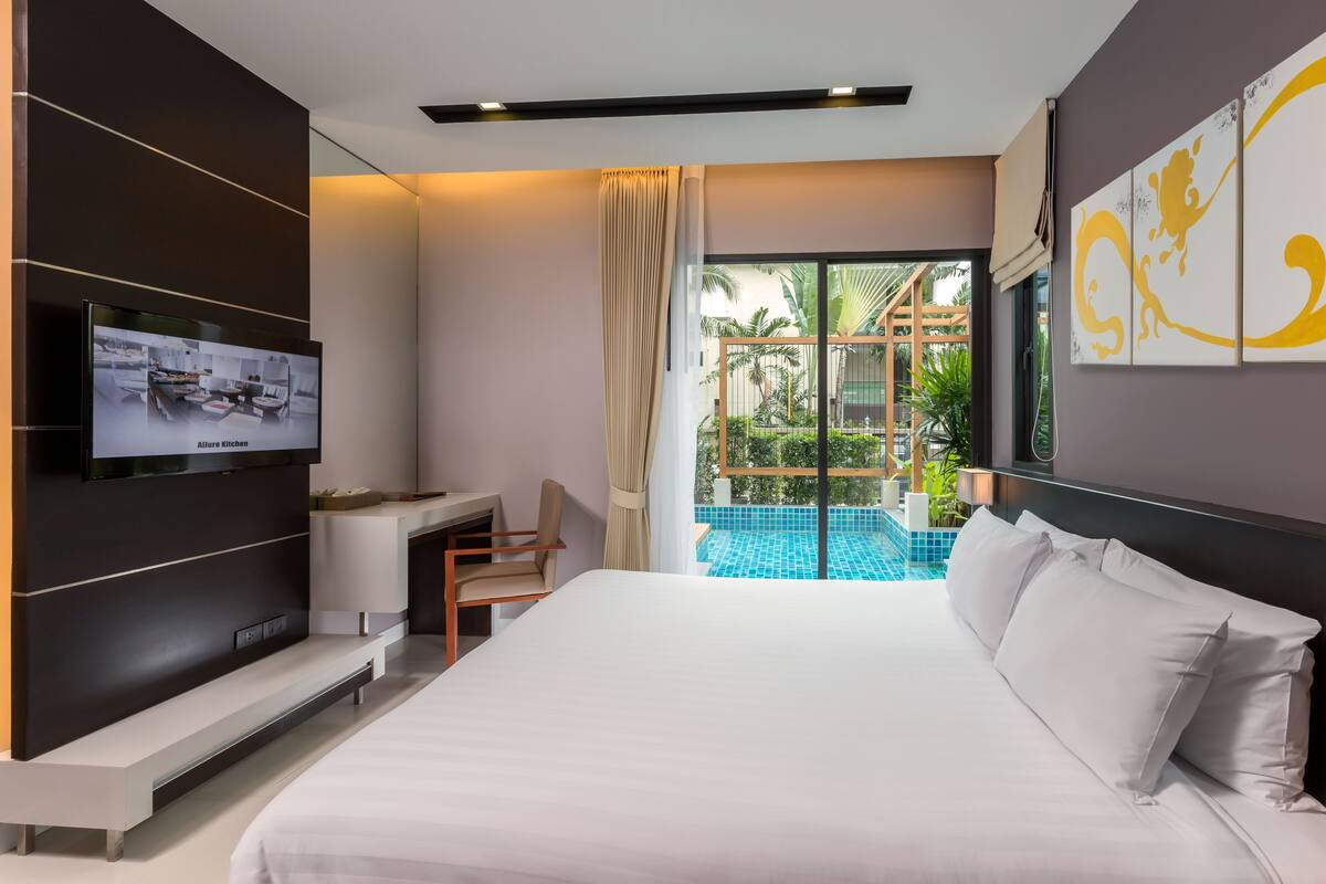 A modern hotel room with a large bed, a TV mounted on the wall, a desk, and a view of an outdoor swimming pool.