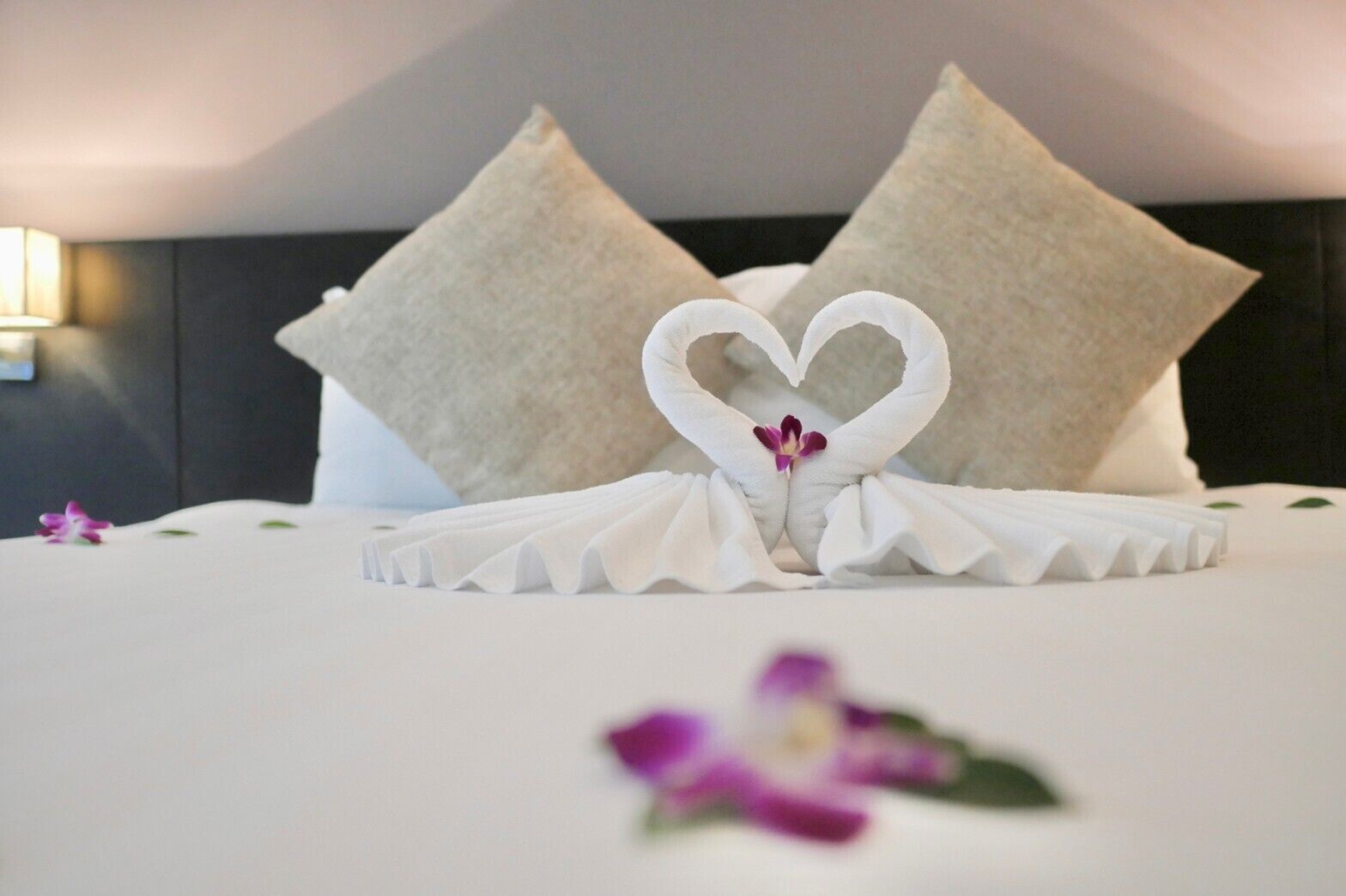 A neatly made bed with a heart-shaped towel arrangement and a single purple flower on the bed.