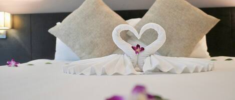 A neatly made bed with a heart-shaped towel arrangement and a single purple flower on the bed.