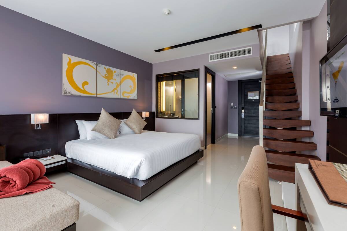 A modern hotel room with a large bed, a desk, a chair, a staircase, and a bathroom visible through an open door.