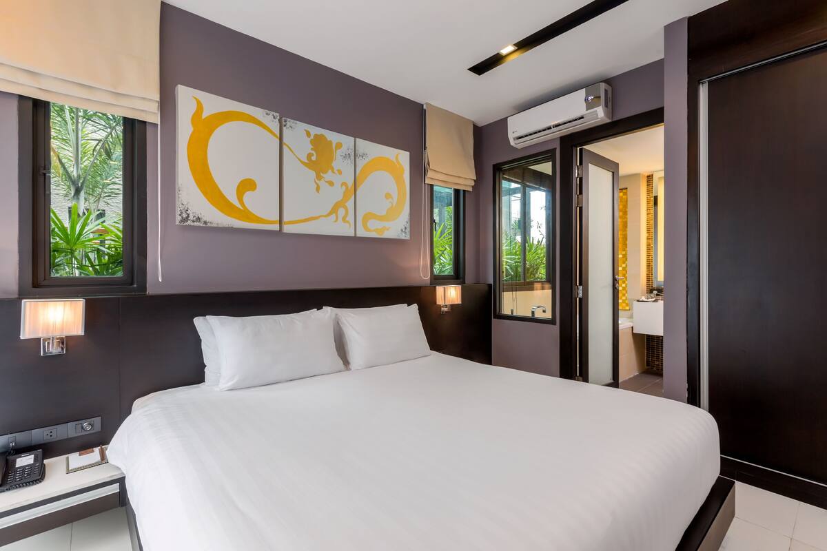 A modern hotel room with a large bed, a bedside table with a phone, a wall-mounted air conditioner, and a window with a view of greenery.