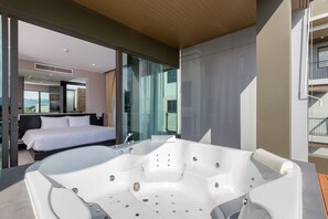 A modern hotel room with a large bathtub, a bed, and a glass wall offering a view of the city.