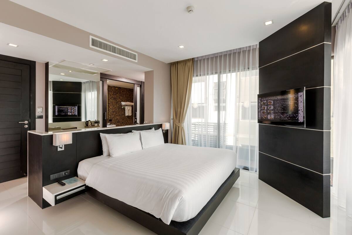 A modern hotel room with a large bed, a flat-screen TV, a built-in desk, and a large window with curtains.