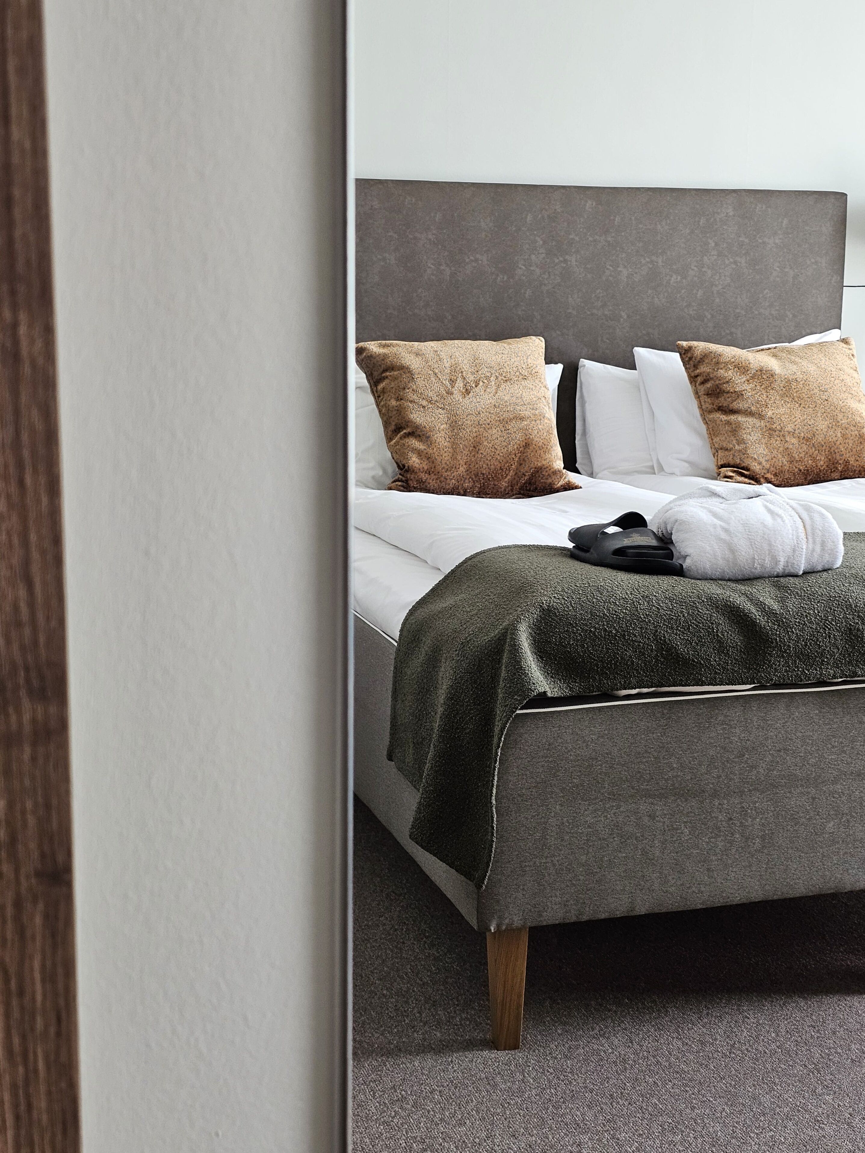 Ein ordentlich bezogenes Bett mit grauem Kopfteil, weißen und braunen Kissen und einer grauen Decke.