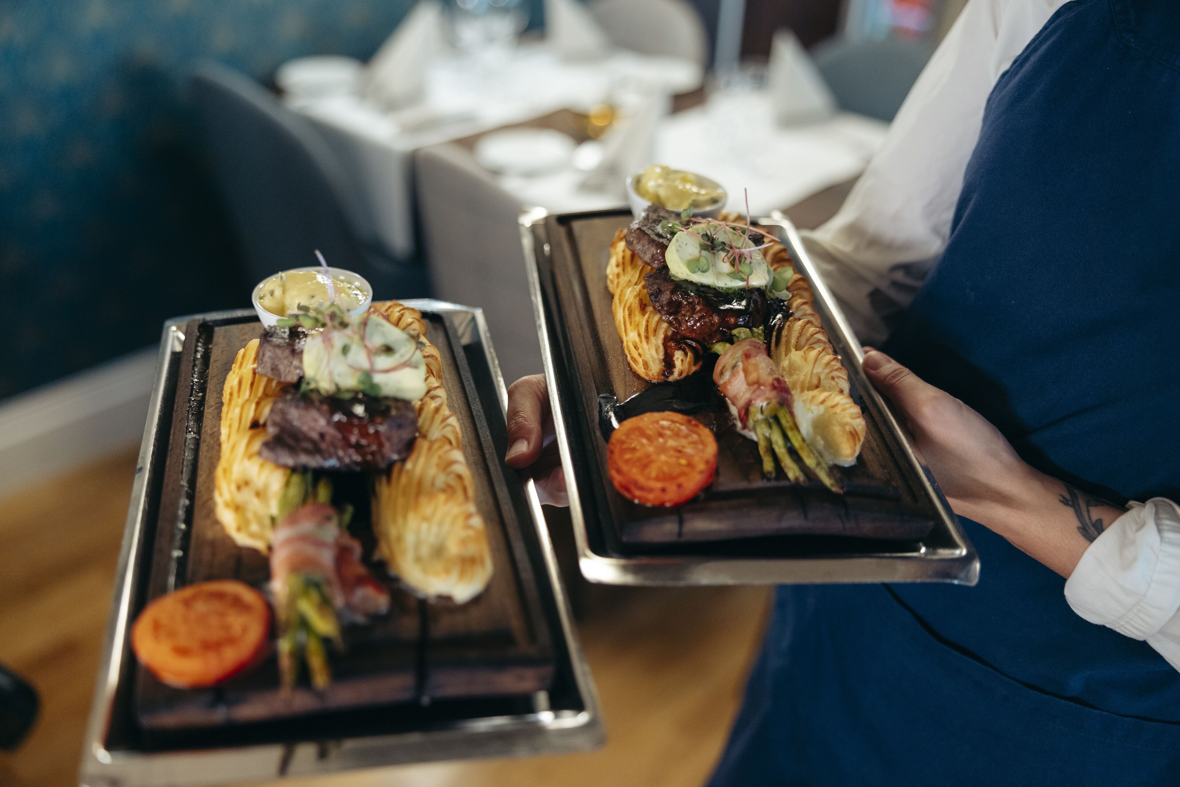 Una persona sosteniendo dos bandejas de comida, con verduras a la parrilla y carne, en un comedor con un sofá azul y un mantel blanco.