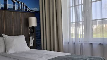 A hotel room with a large bed, a nightstand with a lamp, a view of a bridge, and a window with curtains.