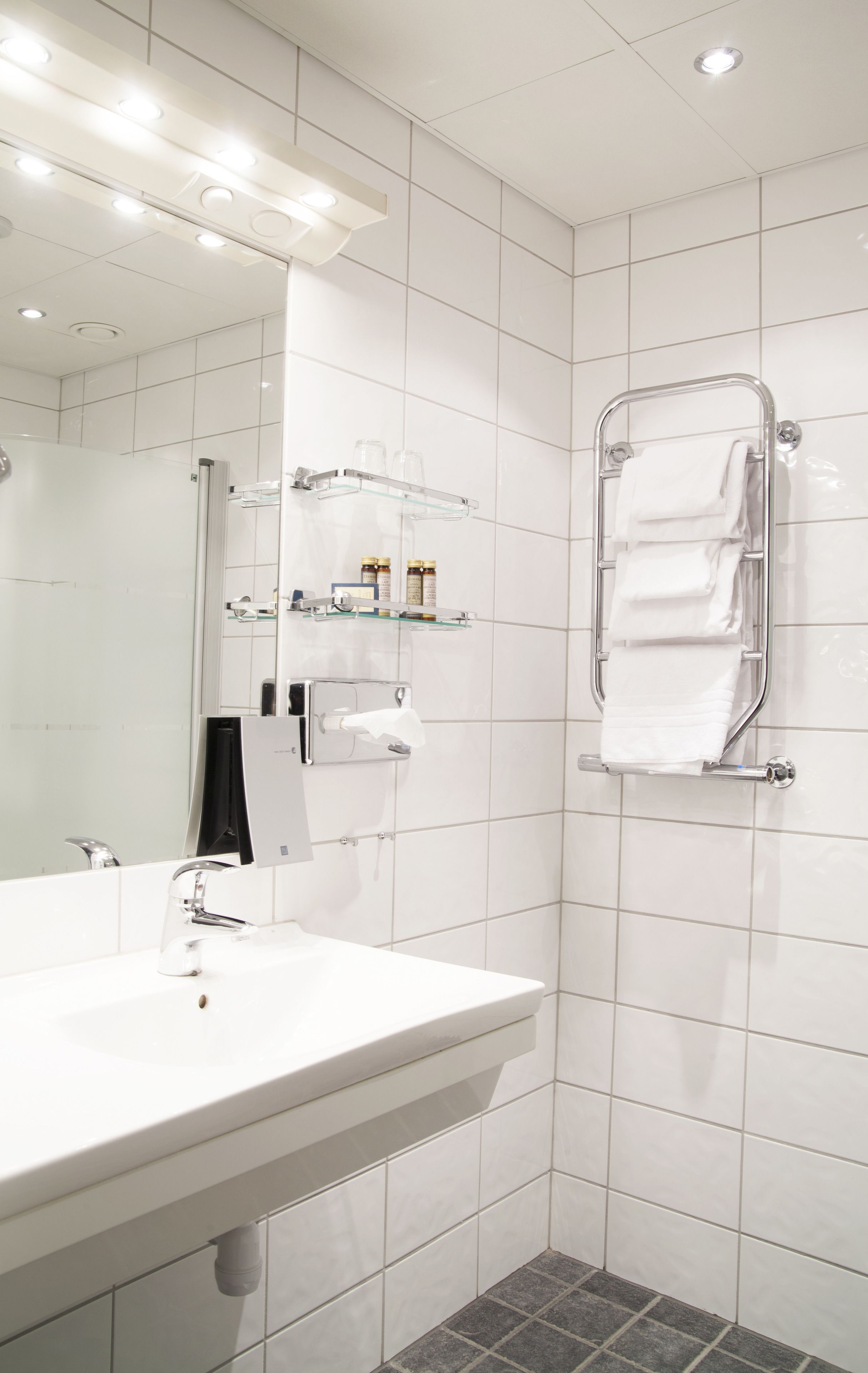 Une salle de bain moderne avec un lavabo blanc, une étagère en verre où sont rangés les articles de toilette, un porte-serviettes et un miroir.