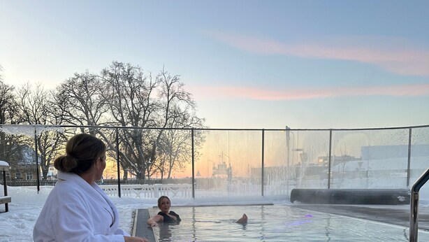 Una persona sentada junto a una piscina exterior climatizada, con una vela y aperitivos, rodeada de nieve y árboles.