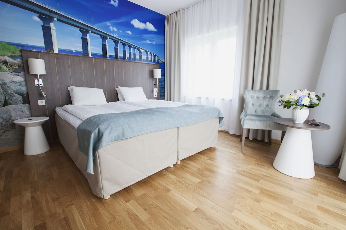 A hotel room with a large bed, a chair, a small table, and a wall mural of a bridge.