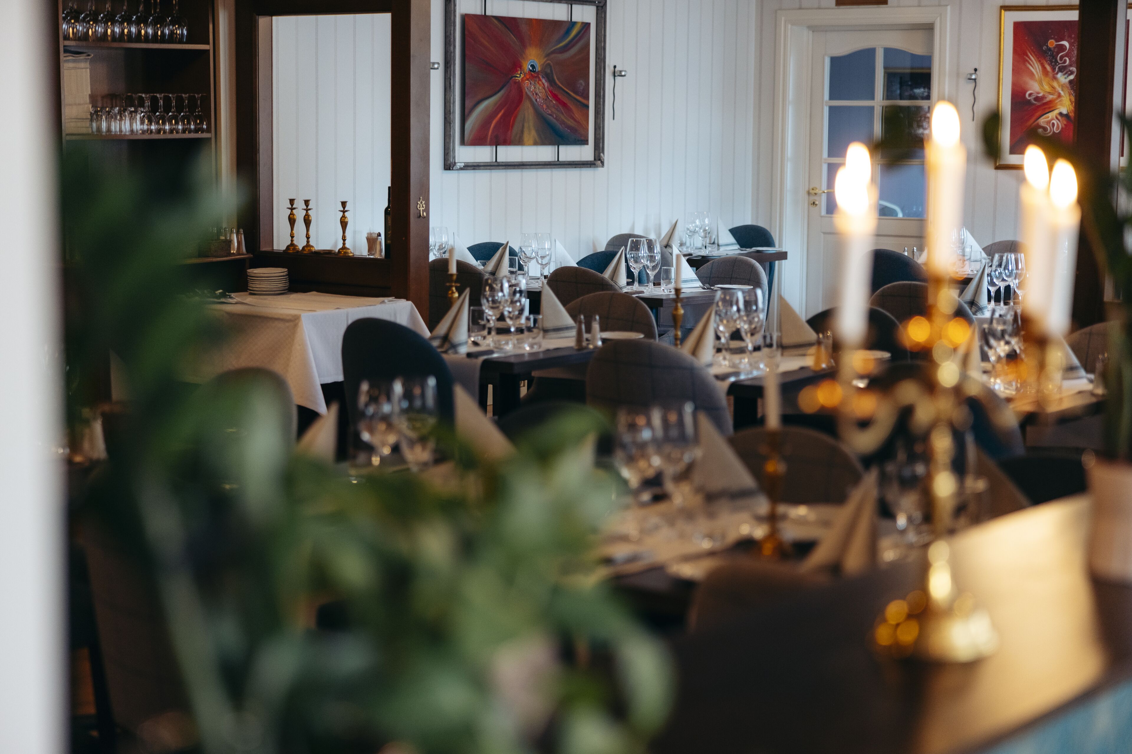 A restaurant with tables set for dining, a wine rack, and a painting on the wall.