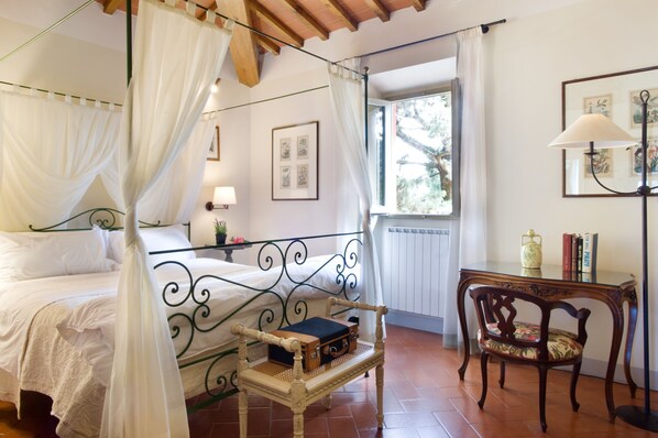 A bedroom with a four-poster bed, a bedside table with a lamp, a chair, and a window with a view of trees. - Casa Villa Rentals by Torre a Cona (Rignano sull'Arno)