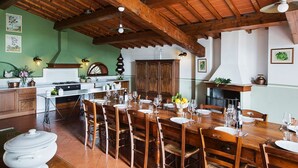 A rustic dining area with wooden tables and chairs, a kitchen in the background, and a high wooden ceiling.