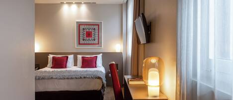 A hotel room with a bed, a desk with a lamp, a TV on the wall, and a window with curtains.