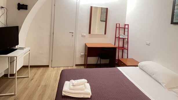 Une petite chambre moderne, équipée d’un lit, d’un bureau, d’une chaise, d’une télévision et d’un miroir.