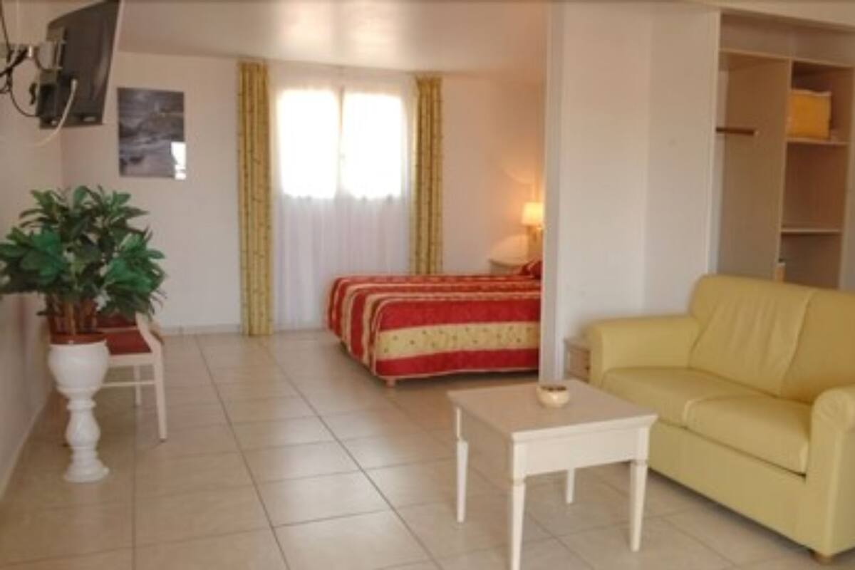 A living room with a yellow sofa, a small white table, a bed with red and white bedding, and a potted plant.