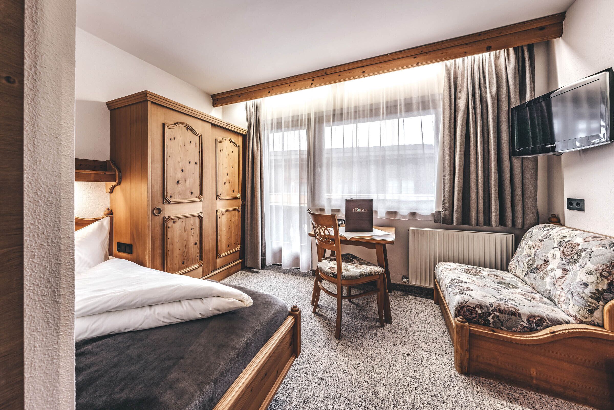 Un dormitorio con una cama, un armario de madera, un escritorio con una silla, un sofá y un televisor montado en la pared.