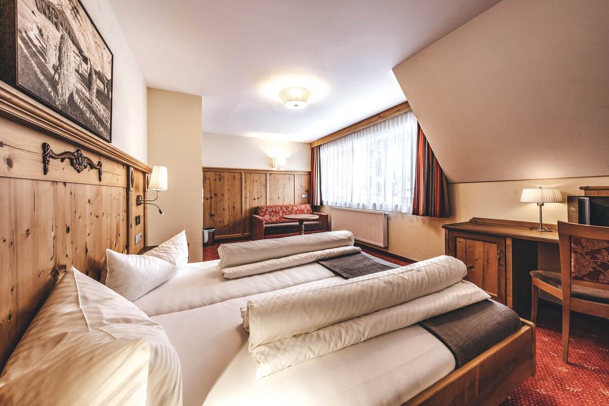 A hotel room with two beds, a desk, a chair, a sofa, and a large window with curtains.