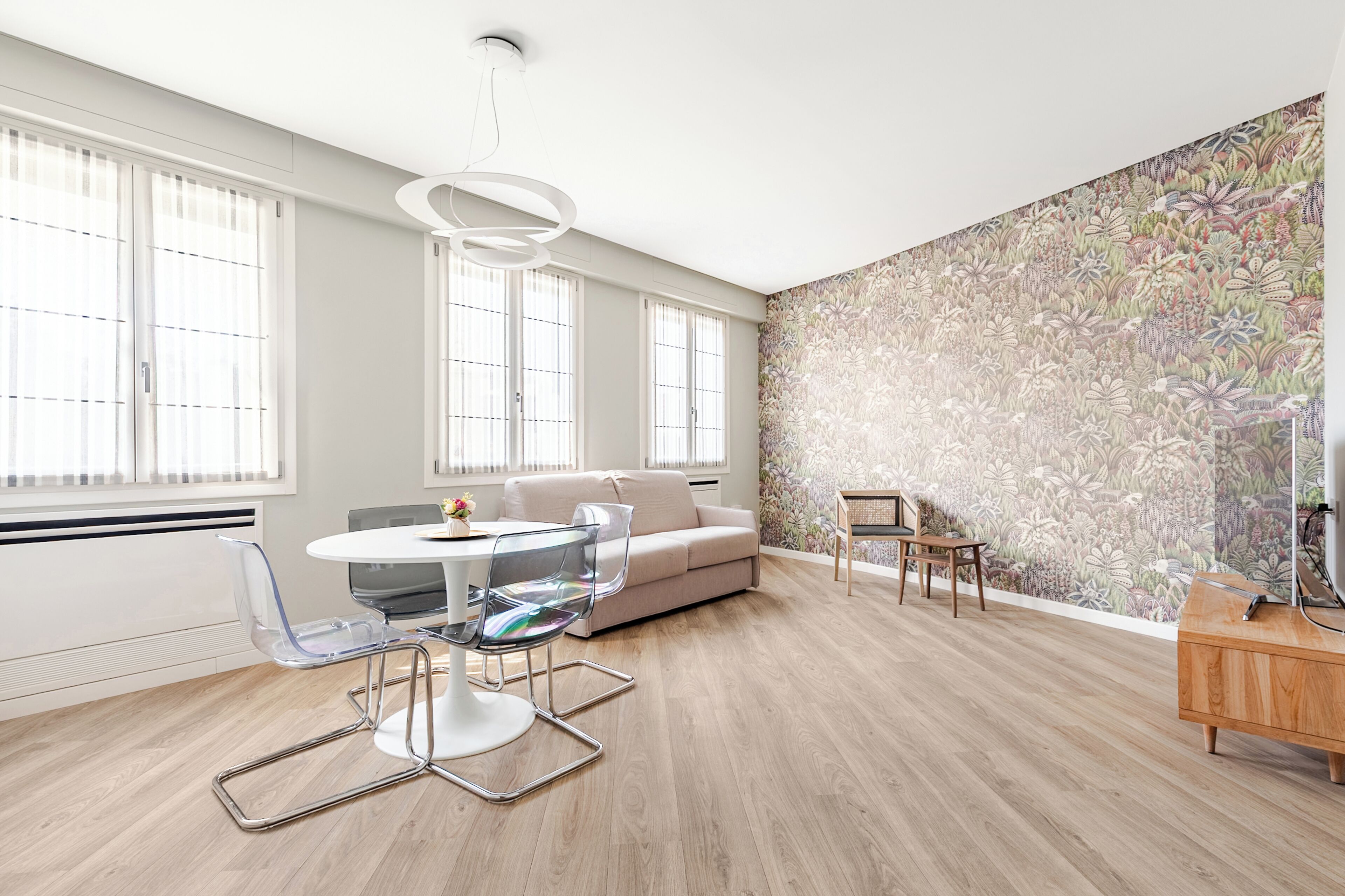 A modern living room with a round glass table, clear chairs, a beige sofa, and a floral wallpaper.