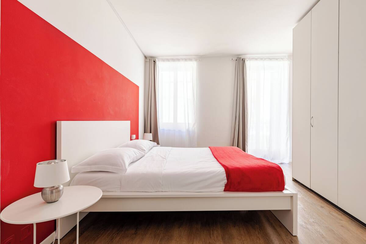 A modern bedroom with a white bed, red accent wall, and a small bedside table with a lamp.