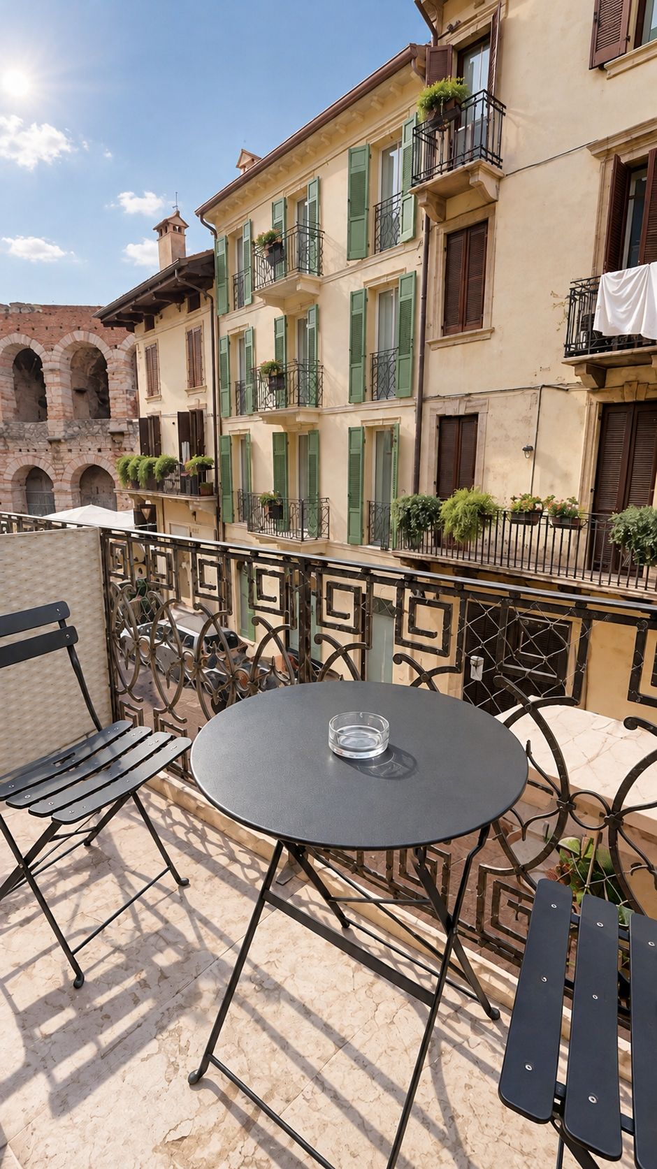 Eine Terrasse im Freien mit einem runden Tisch und Stühlen, einem schmiedeeisernen Geländer und Gebäuden mit grünen Fensterläden im Hintergrund.