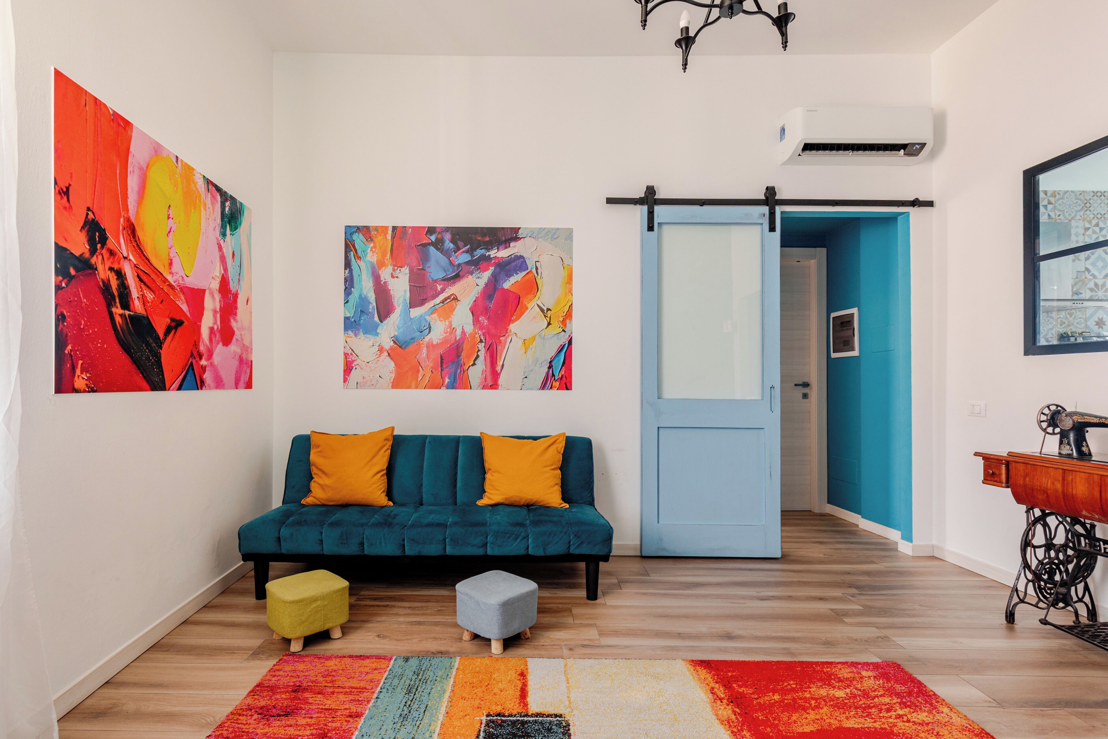A modern living room with a teal sofa, colorful abstract art, a sliding door, and a vintage sewing machine.