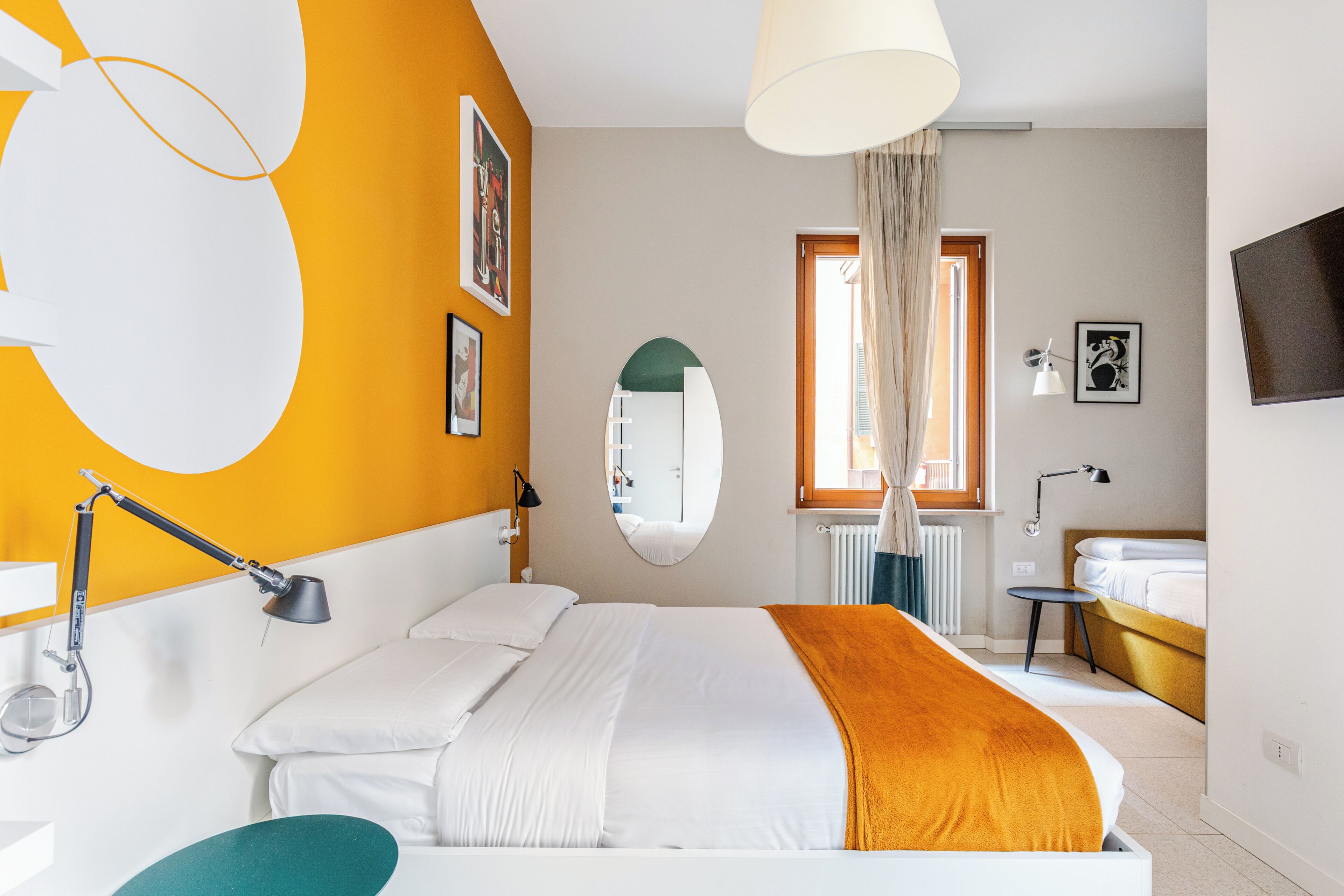 A modern hotel room with a bed, a desk, a chair, a desk lamp, a mirror, a window with curtains, a radiator, a TV, and artwork on the walls.
