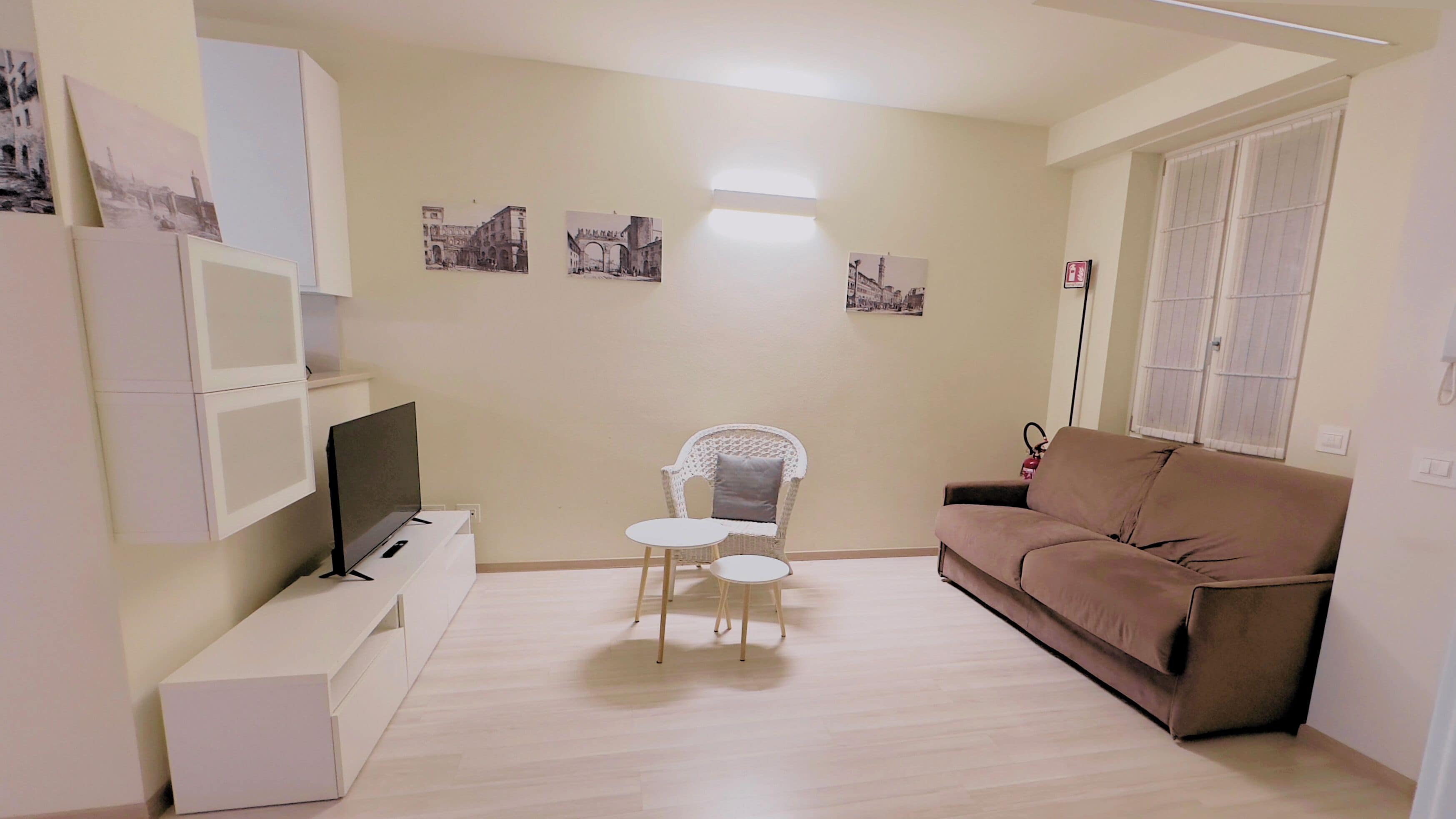 A living room with a flat-screen TV, a brown sofa, a white chair, a small table, and wall-mounted pictures.