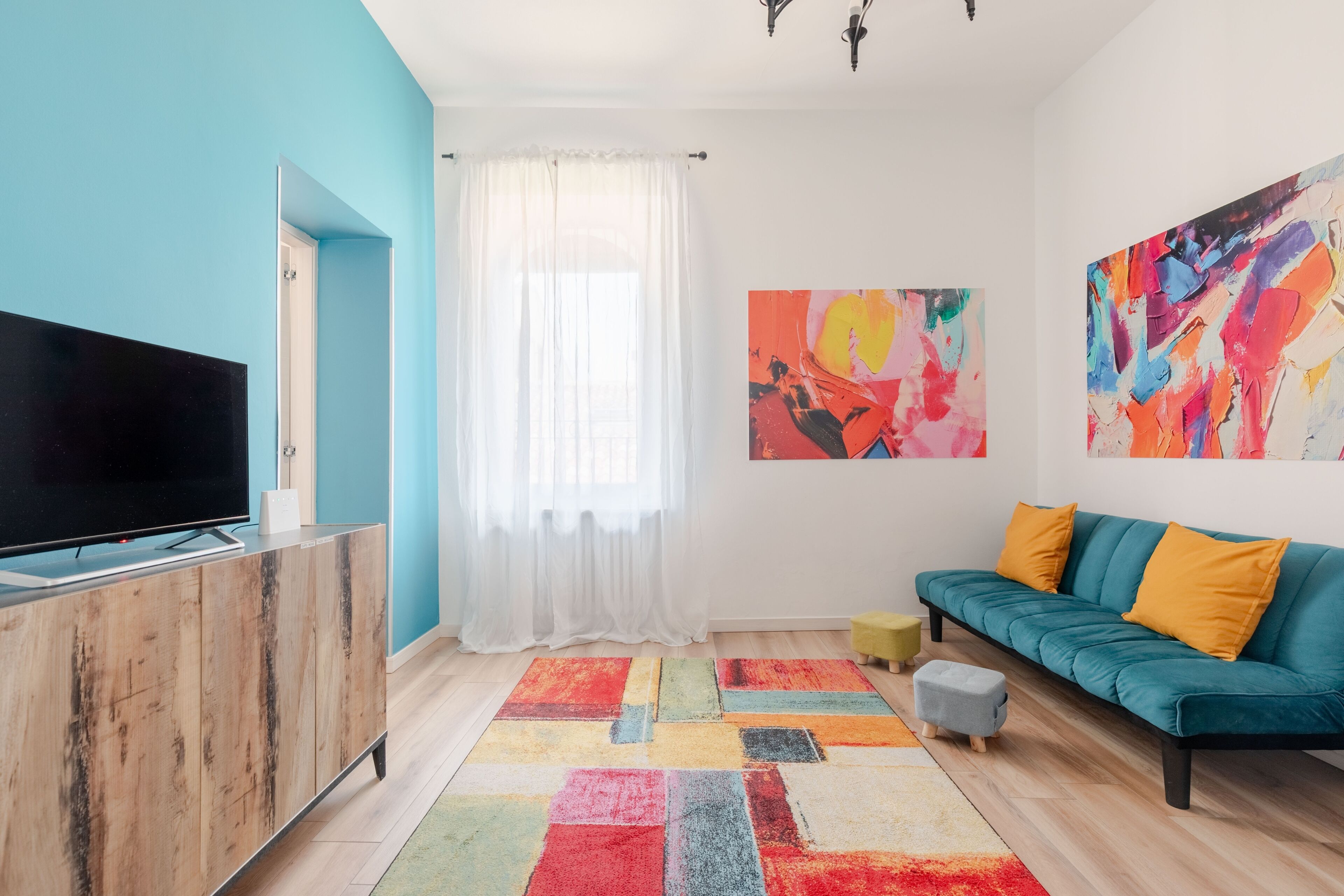 A modern living room with a flat-screen TV, a teal sofa, colorful rugs, and abstract wall art.