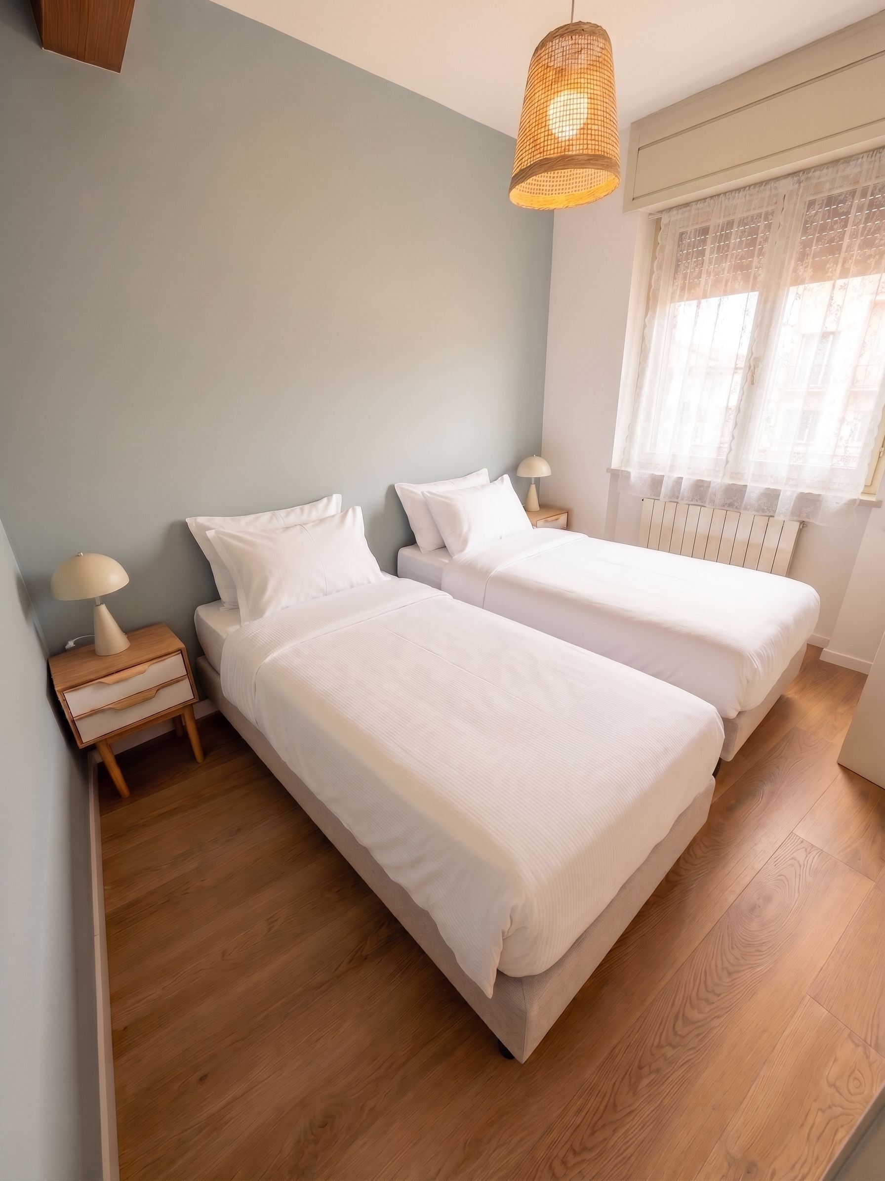 Un dormitorio con dos camas, una mesita de noche con lámpara, una ventana con cortinas traslúcidas y una lámpara colgante.